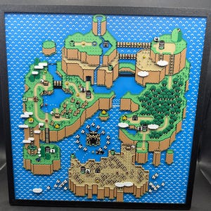 May include: A framed, three-dimensional artwork depicting a colorful map of a video game world. The map features brown landmasses, blue water, green trees, and various game elements. The artwork is encased in a black frame.
