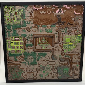 May include: A framed, three-dimensional map with a detailed, textured design. The map features various colors, including shades of brown, green, and pink, with a black frame. The map appears to depict a fantasy landscape with buildings and pathways.