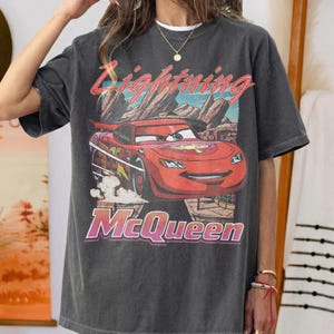 May include: Dark gray t-shirt featuring a graphic of Lightning McQueen from the Cars movie. The design includes the text "Lightning McQueen" in a retro style, with a red race car against a desert landscape.