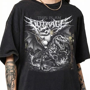 May include: Dark gray t-shirt featuring a graphic of a cat riding a dinosaur, holding a sword, with the text "THIS IS AN OUTRAGE" in a stylized font. The design is in black and white.
