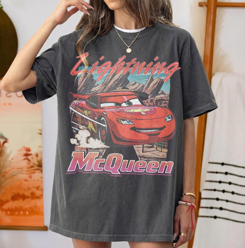 May include: Dark gray t-shirt featuring a graphic of Lightning McQueen from the Cars movie. The design includes the text "Lightning McQueen" in a retro style, with a red race car against a desert landscape.