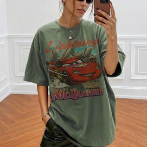 May include: Olive green t-shirt featuring a graphic of Lightning McQueen from the Cars movie. The design includes the text "Lightning McQueen" in red and pink. The shirt has short sleeves and a relaxed fit.