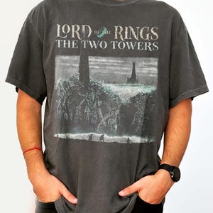 Puede incluir: Camiseta gris oscuro con el texto "LORD of the RINGS THE TWO TOWERS" y un gráfico de dos torres y figuras. La camiseta tiene un aspecto vintage y descolorido.