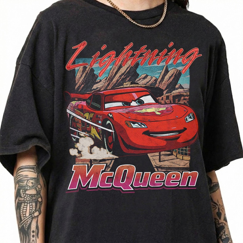 May include: Dark gray t-shirt featuring a graphic of Lightning McQueen from the movie Cars. The design includes the text "Lightning McQueen" in red and "McQueen" in pink, with a backdrop of a desert landscape.