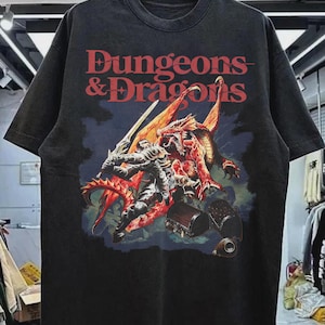 May include: Black t-shirt featuring a graphic of a knight battling a red dragon, with the words "Dungeons & Dragons" in red lettering. The design is set against a dark blue background. The shirt is hanging on a black hanger.