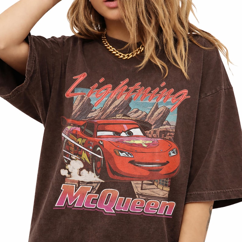 May include: Brown t-shirt featuring a graphic of Lightning McQueen from the movie Cars. The design includes the text "Lightning McQueen" and "McQueen" in a stylized font, with a desert landscape background.