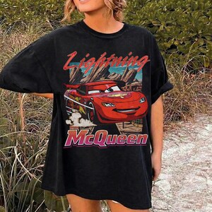 May include: Black t-shirt featuring a graphic of Lightning McQueen from the Cars movie. The design includes the text "Lightning McQueen" in red and "McQueen" in pink. The shirt has a vintage, distressed look.