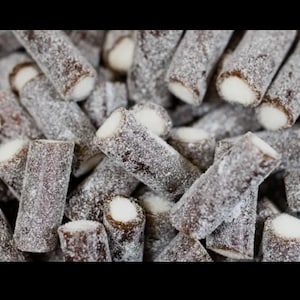 Può includere: Primo piano di un mucchio di caramelle cilindriche. Le caramelle sono marrone scuro con un centro bianco e sono ricoperte da una sostanza zuccherina bianca. Le caramelle sono disposte in modo casuale, riempiendo l'inquadratura.
