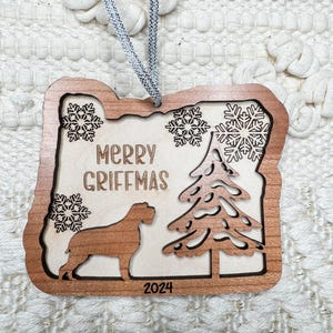 May include: Wooden Christmas ornament with a light brown frame and a light beige interior. The ornament features a dog silhouette, snowflakes, a Christmas tree, and the text "MERRY GRIFFMAS 2024". A silver and white string is attached for hanging.