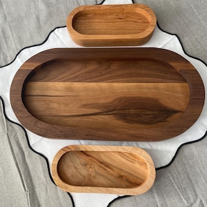 Puede incluir: Tres bandejas ovaladas de madera de diferentes tamaños. La bandeja más grande es de color marrón oscuro, la mediana es de color marrón claro y la más pequeña es de color beige claro. Las bandejas están sobre un paño blanco con borde festoneado negro.