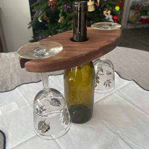 May include: A wooden wine glass and bottle holder with a dark brown finish. The holder has two circular cutouts for wine glasses and a central hole for a wine bottle. Two wine glasses with leaf designs are hanging from the holder.