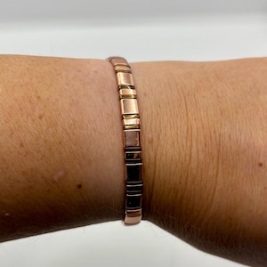 Puede incluir: Una pulsera de color cobre con eslabones rectangulares, con secciones alternas de cobre, oro y negro. La pulsera se lleva en la muñeca, mostrando su diseño segmentado y brillo metálico.