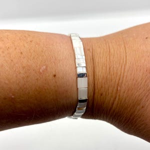 Könnte beinhalten: Ein silbernes Armband mit rechteckigen weißen und silbernen Perlen. Das Armband wird an einem Handgelenk getragen, wodurch sein Design und der Farbkontrast hervorgehoben werden. Das Design des Armbands ist schlicht und elegant.