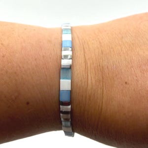 Op de afbeelding: Een armband met vierkante kralen in lichtblauw, wit en zilver. De armband wordt om een pols gedragen en lijkt te zijn gemaakt van kleine, vierkante kralen die aan elkaar zijn geregen. Een delicaat sieraad.