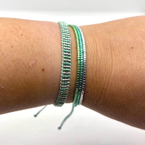 Könnte beinhalten: Zwei Armbänder aus Perlen in Grün- und Silbertönen werden an einem Handgelenk gezeigt. Die Armbänder bestehen aus kleinen, rechteckigen Perlen, die miteinander verwoben sind. Ein Armband ist breiter als das andere. Die Armbänder haben verstellbare Bänder.