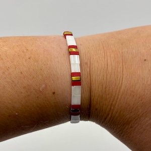 Op de afbeelding: Een armband met een patroon van witte, rode en gouden rechthoekige kralen. De armband wordt om een pols gedragen, waarbij de afwisselende kleuren en het algehele ontwerp worden getoond. De kralen zijn aan elkaar geregen, waardoor een flexibel en stijlvol accessoire ontstaat.
