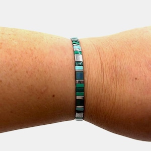 May include: A bracelet featuring rectangular beads in silver, turquoise, green, and dark green. The bracelet is worn on a wrist and has a modern design. The small, square beads create a geometric pattern. The bracelet is approximately 18 cm in length.