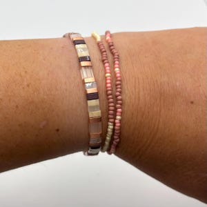 Op de afbeelding: Drie armbanden worden op een pols getoond. Eén armband is gemaakt van rechthoekige kralen in bruin-, beige- en roségoudtinten. De andere twee armbanden zijn gemaakt van kleine, ronde kralen in roze en crèmekleur.