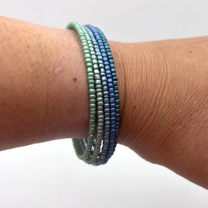 May include: A multi-strand bracelet composed of small, round, glossy beads in a gradient color scheme. The beads transition from a light mint green to a medium sage, then to a soft teal, and finally to a deep navy blue, creating a cohesive look.