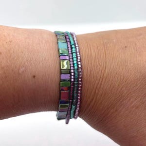 May include: A stack of four bracelets featuring one strand of flat, rectangular, iridescent tiles in shades of blue, green, and purple, alongside three thinner strands of small, round, metallic beads in purple, teal, and light purple hues.
