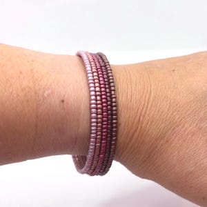 Peut inclure: Un bracelet multi-rangs composé de petites perles rondes dans un dégradé de couleurs allant du rose clair au mauve foncé. Les perles présentent un léger éclat métallique et sont disposées en cinq rangées distinctes autour du poignet.