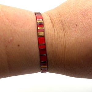 Op de afbeelding: Een rode en gouden armband met rechthoekige kralen. De armband wordt om een pols gedragen en heeft afwisselend rode en goudkleurige segmenten. Het is een sieraad.