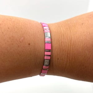 Könnte beinhalten: Ein rosa-silbernes Armband aus kleinen, quadratischen Perlen. Das Armband hat abwechselnd Abschnitte aus leuchtend rosa, hellrosa und silberfarbenen Perlen. Das Armband wird am Handgelenk getragen.