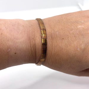 May include: A metallic bronze-colored stretch bracelet composed of small, rectangular, polished segments. The bracelet features a repeating pattern of smooth, flat links that create a flexible, continuous band around the wrist.