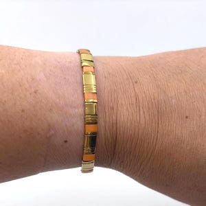 May include: A stretch bracelet featuring alternating rectangular beads in a solid orange color and polished gold-tone metal segments. The gold-tone segments include both smooth surfaces and sections with fine horizontal etched line detailing.