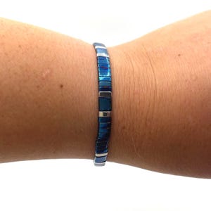 May include: A blue bracelet with rectangular, metallic beads. The bracelet is worn on a wrist, showcasing its colour and design. The beads are a deep blue with silver accents, creating a modern and stylish look. The bracelet is a piece of jewellery.