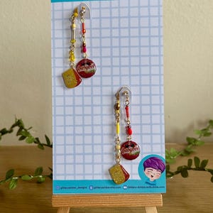 Könnte beinhalten: Handgefertigte Ohrringe mit einem skurrilen Design. Jeder Ohrring hat eine goldene Kette mit bunten Perlen, einen roten Babybel-Käse-Anhänger und einen gelben Käse-Keil-Anhänger. Der Hintergrund ist ein blau-weißes Gittermuster.