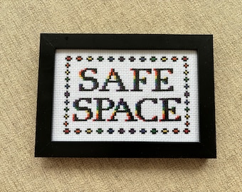 May include: A framed cross-stitch with the words "SAFE SPACE" in rainbow colors. The letters are outlined in black and the background is white.