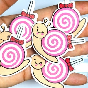 Könnte beinhalten: Eine Sammlung skurriler Aufkleber in Schneckenform. Jeder Aufkleber zeigt eine rosa und weiß wirbelnde Lutscher-Schale, ein lächelndes Gesicht und eine rote Schleife. Die Aufkleber werden in einer Hand gehalten.
