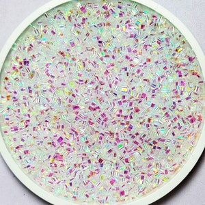 May include: A white round container filled with iridescent glitter. The glitter is a mix of small squares and rectangles with a rainbow sheen.