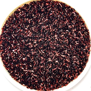 Puede incluir: Una imagen de primer plano de un cuenco lleno de pequeñas cuentas metálicas rojas. Las cuentas son brillantes y tienen un acabado ligeramente iridiscente.