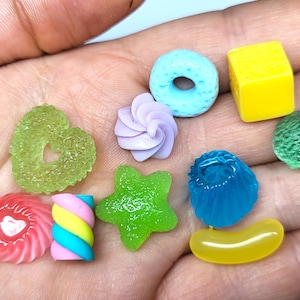 Può includere: Una collezione di ciondoli per alimenti in miniatura colorati, tra cui un cuore verde, un vortice rosa e bianco, una stella verde, un ciambellone blu, un fagiolo di gelatina giallo, un vortice verde e bianco, un cupcake blu, un cubo giallo e una sfera verde irregolare.