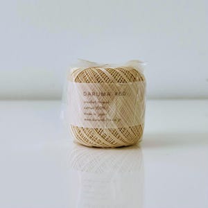May include: A spool of Daruma #60 crochet thread, made of 100% cotton, is packaged in clear plastic. The label displays the brand name and product details. The thread is a light beige color, suitable for various crafting projects.
