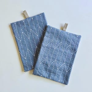 May include: Two rectangular, slate blue fabric pot holders with white zigzag stitching. Each pot holder has a small, tan loop at the top for hanging. The pot holders are made of a textured fabric.