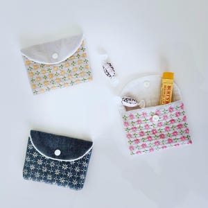 May include: Three small, handmade fabric pouches with button closures. Two pouches feature floral embroidery in yellow and pink, while the third is navy blue with white starburst patterns. One pouch contains a lip balm.