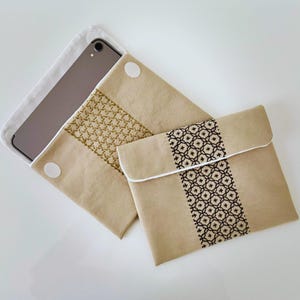 May include: Beige fabric tablet sleeve with a decorative embroidered panel. One sleeve has a gold geometric pattern, the other has a black and white floral design. A tablet is partially visible inside one sleeve, secured with a white velcro closure.