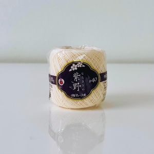 May include: A spool of off-white thread, wrapped in clear plastic, with a label featuring Japanese text and a floral design. The label is dark purple with gold accents. The thread is labeled as #40.