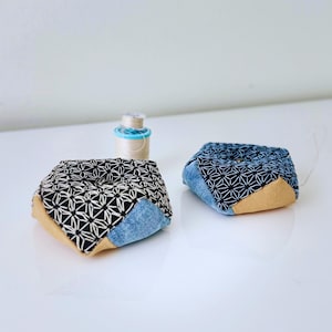 May include: Two handmade pincushions with geometric fabric patterns. One pincushion features a black and white design, while the other has a blue and white pattern. Both have denim and tan fabric accents. A spool of thread sits in the background.
