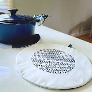 May include: A blue enameled cast iron pot with a lid and a white and navy blue patterned pot holder. A white round trivet with a navy blue geometric pattern is in the foreground. Kitchen and dining.