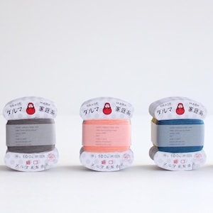 May include: A row of colorful thread spools with Japanese text and a Daruma doll logo. The spools are in shades of pink, gray, orange, blue, and black. Each spool has a label with text and a small red Daruma doll illustration.