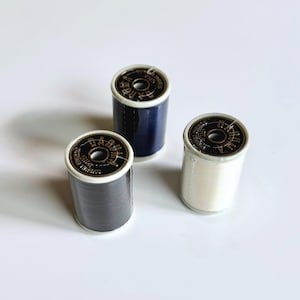 May include: Three spools of sewing thread in navy blue, gray, and white. Each spool has a black and gold label with text. The spools are cylindrical and sit on a white surface. The thread is tightly wound.