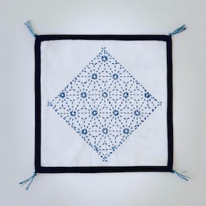 May include: Square white cushion with a navy blue border and tassels at each corner. The center features a diamond-shaped pattern of blue embroidered floral designs. A decorative textile item.