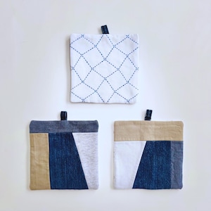 May include: Three square fabric pot holders with hanging loops. The top one is white with a blue stitched diamond pattern. The bottom two are patchwork designs featuring panels of tan, dark blue denim, light gray, and white fabric segments.