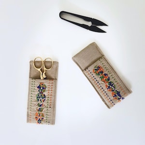 May include: An overhead shot shows a pair of gold scissors in a tan fabric case with colorful geometric embroidery. A second case and a pair of black snips are also visible. The items are set against a white background, suggesting a craft or sewing theme.