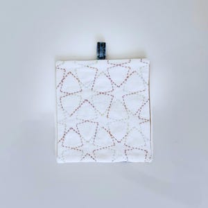 May include: Square white fabric item with a repeating triangle pattern in brown and green stitching. A small, dark blue fabric loop is attached to the top edge. The item is likely a coaster or small mat.
