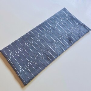 May include: A rectangular, gray fabric item with a white zigzag stitch pattern. The item appears to be a table runner or placemat, with a simple, minimalist design. The fabric has a slightly textured appearance.